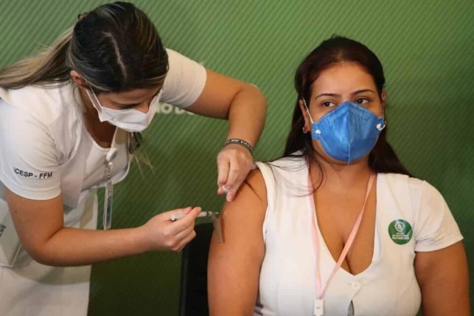 Mulher profissional de saúde de pé aplica vacina contra covid-19 no braço de outra profissional de saúde que está sentada. Ambas estão de máscara