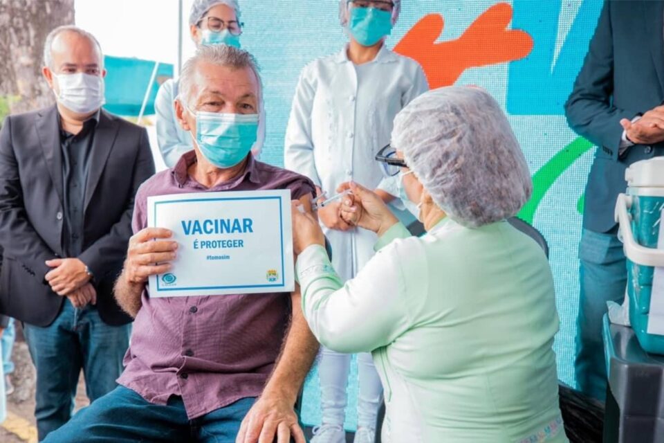 Idoso sentado sendo vacinado contra a covid-19 por enfermeira. Ele segura uma placa com a inscrição "vacinar é proteger". Todos estão de máscara.