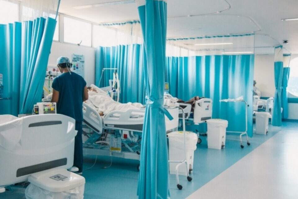 Profissional de saúde próximo a leito de paciente com covid-19. Há cortinas azuis e outros leitos compondo a imagem, no Hospital Metropolitano em Maceió