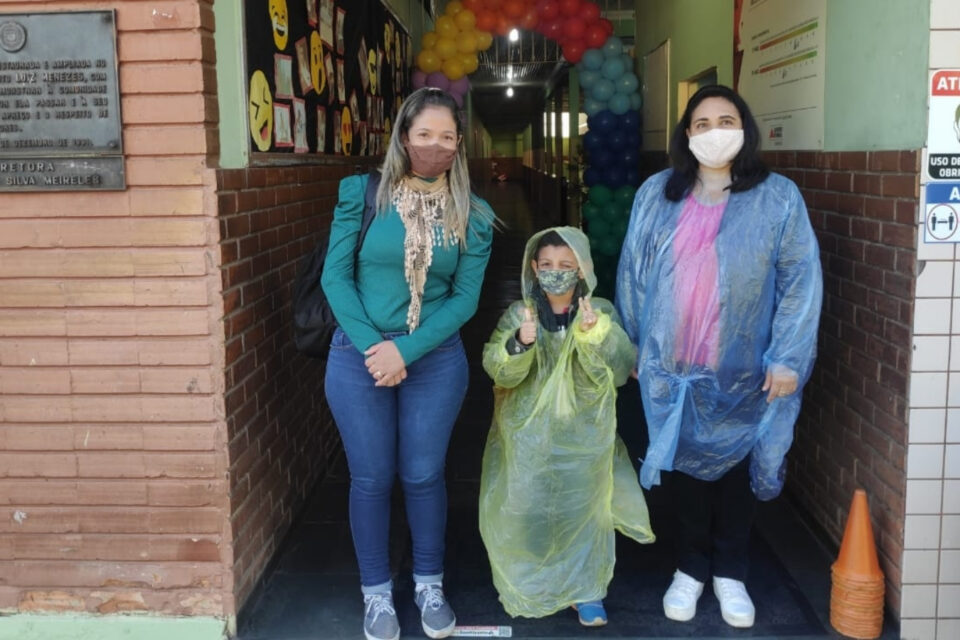 Aluna de capa de chuva amarela entre duas mulheres em uma escola. Todas estão de máscara e a mulher da direita usa uma proteção descartável contra contaminação, sobre a roupa.