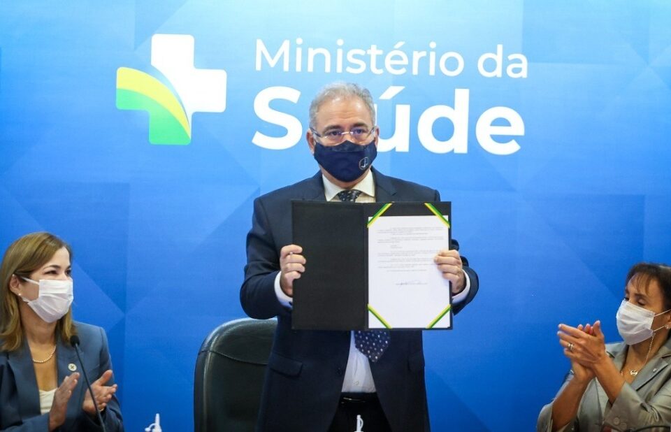 Ministro da Saúde, Marcelo Queiroga, usa terno e máscara azul e está de pé expondo um documento. Duas mulheres sentadas e de máscaras aplaudem usando máscaras brancas. Ao fundo um cartaz azul com nome e marca do Ministério da Saúde