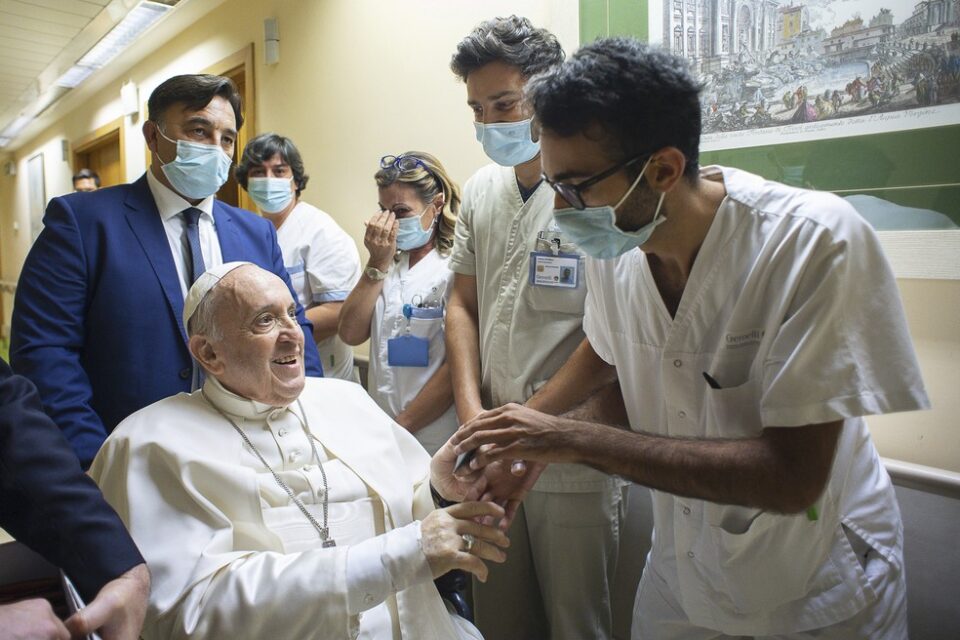 Papa sentado na cadeira de rodas com enfermeiros segurando sua mão