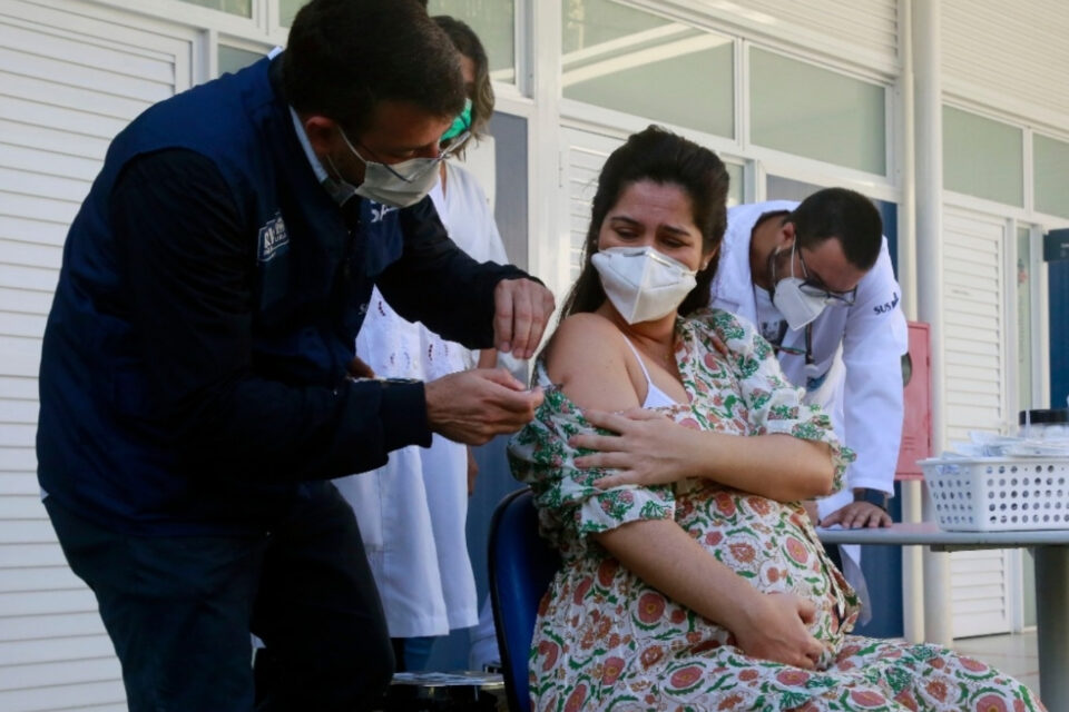 Grávida de máscara, sentada, sendo vacinada com dose da vacina Pfizer contra a covid-19