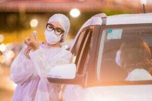 Enfermeira de jaleco, máscara e touca brancas, prepara dose de vacina para aplicar em mulher que espera dentro de carro.