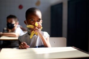 Menino come uma banana em sala de aula. Ele está com máscara abaixada e tem um colega de máscara na carteira escolar ao fundo