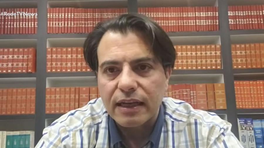 Homem de camisa xadrez sentado à frente de estante de livros