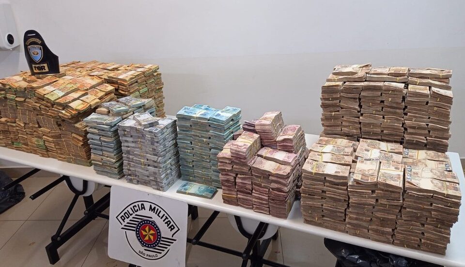 Notas de dinheiro empilhadas sobre mesa com placa da Polícia Militar